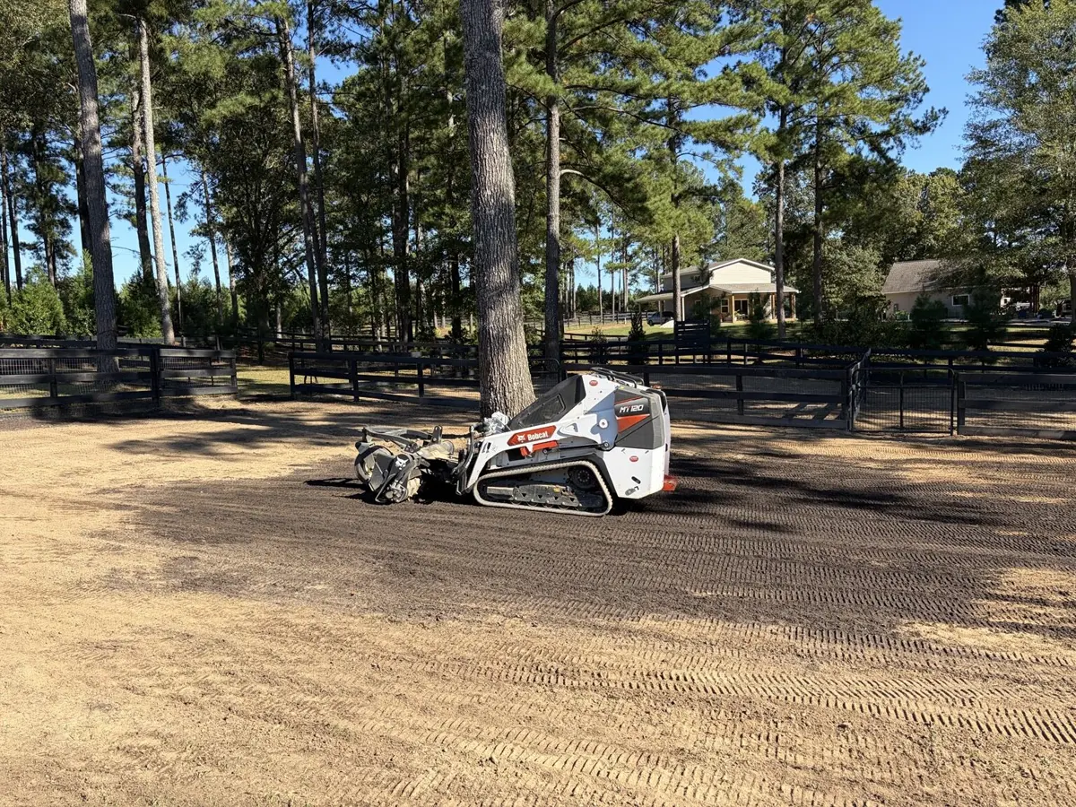 RSL compact track loader grading a large dirt area beside a fenced pasture and pine trees